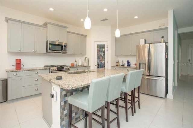 kitchen featuring an island with sink, sink, hanging light fixtures, stainless steel appliances, and light stone countertops