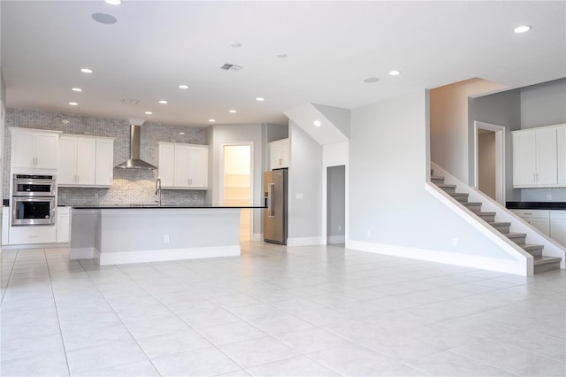 kitchen featuring white cabinetry, a center island with sink, wall chimney range hood, stainless steel appliances, and backsplash