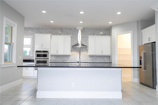 kitchen featuring appliances with stainless steel finishes, wall chimney exhaust hood, white cabinetry, decorative backsplash, and a kitchen island with sink