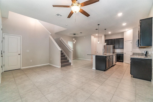 kitchen with an island with sink, ceiling fan, stainless steel refrigerator, pendant lighting, and light stone counters