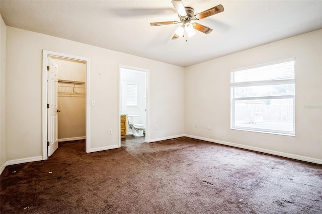 unfurnished bedroom featuring a closet, ensuite bath, dark carpet, ceiling fan, and a walk in closet