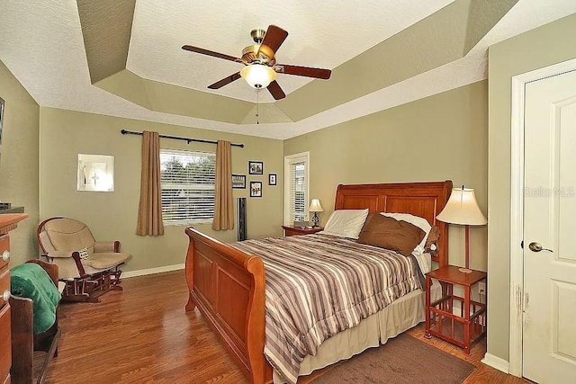 bedroom featuring a ceiling fan, wood finished floors, baseboards, a tray ceiling, and a textured ceiling