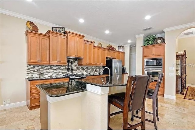 kitchen with backsplash, dark stone countertops, crown molding, appliances with stainless steel finishes, and an island with sink