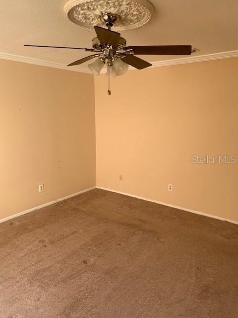 empty room with a ceiling fan, dark colored carpet, crown molding, and baseboards