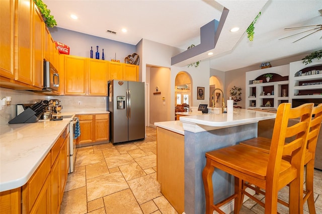 kitchen with ceiling fan, backsplash, a kitchen island with sink, appliances with stainless steel finishes, and a kitchen breakfast bar