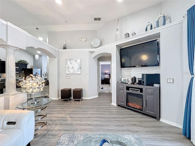 living room featuring light wood-type flooring, a fireplace, a towering ceiling, and decorative columns