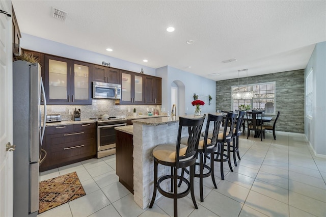 kitchen with light tile patterned floors, appliances with stainless steel finishes, light stone counters, an island with sink, and a kitchen bar