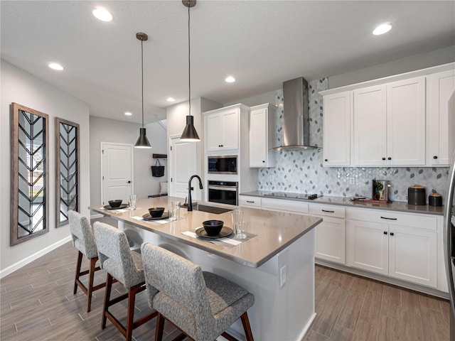 kitchen with a kitchen island with sink, hanging light fixtures, wall chimney range hood, white cabinets, and black appliances