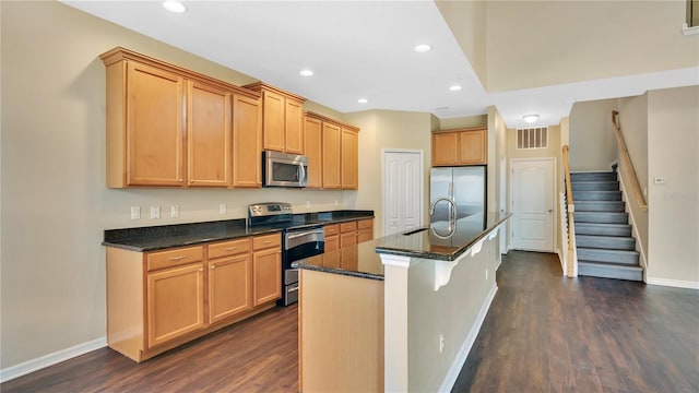 kitchen with appliances with stainless steel finishes, sink, dark wood-type flooring, dark stone counters, and a center island with sink