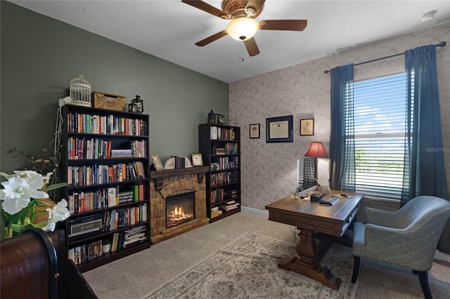 carpeted home office featuring a stone fireplace and ceiling fan