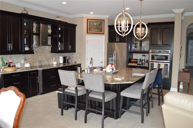 kitchen with a center island with sink, an inviting chandelier, light stone countertops, stainless steel appliances, and wall chimney exhaust hood