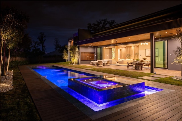 pool at night featuring ceiling fan, an outdoor hangout area, a wooden deck, and an in ground hot tub