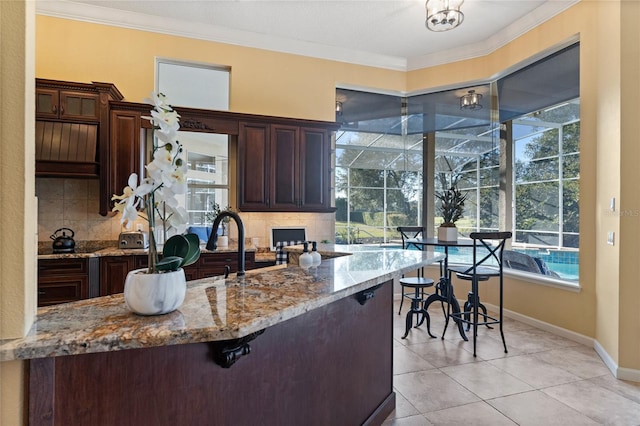 kitchen with light tile patterned floors, dark brown cabinetry, light stone counters, ornamental molding, and decorative backsplash