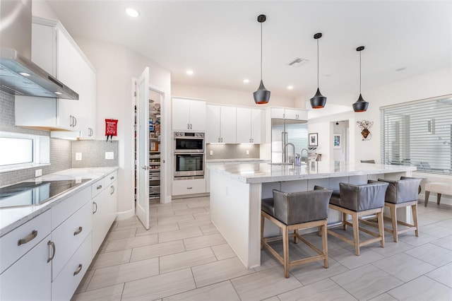 kitchen featuring decorative light fixtures, wall chimney range hood, white cabinetry, an island with sink, and a breakfast bar