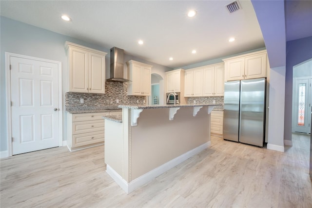 kitchen featuring light hardwood / wood-style floors, an island with sink, stainless steel appliances, light stone counters, and wall chimney exhaust hood