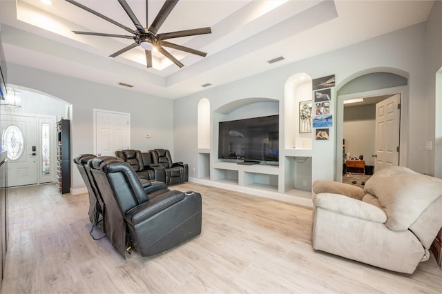 living room featuring built in shelves, light wood-type flooring, ceiling fan, and a tray ceiling