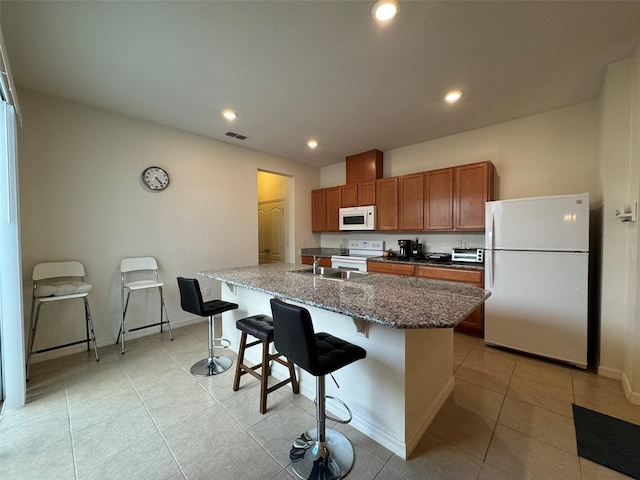 kitchen with a breakfast bar area, dark stone counters, light tile patterned floors, a center island with sink, and white appliances