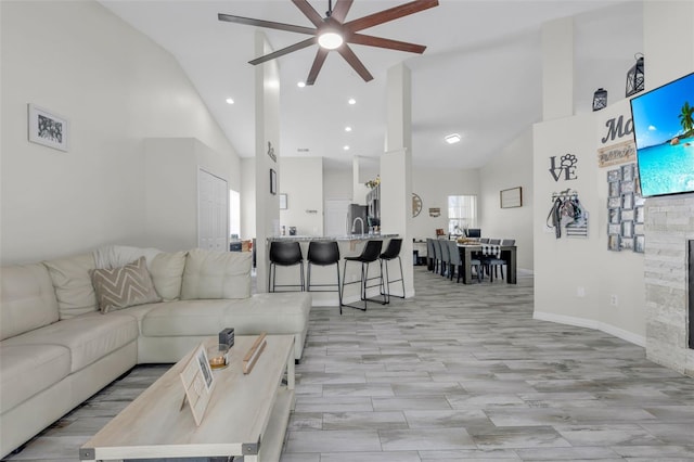 living room featuring ceiling fan, sink, a stone fireplace, and high vaulted ceiling