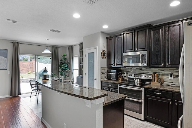 kitchen featuring stainless steel appliances, backsplash, dark stone counters, and an island with sink