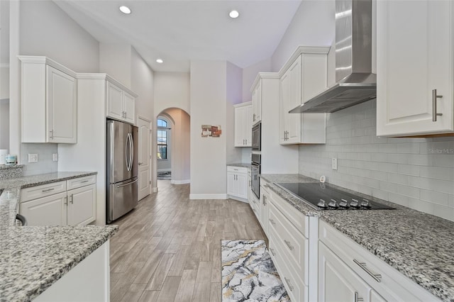 kitchen featuring white cabinets, light stone counters, stainless steel appliances, wall chimney range hood, and light hardwood / wood-style flooring