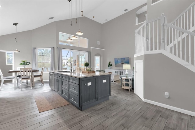 kitchen featuring an island with sink, decorative light fixtures, high vaulted ceiling, light stone counters, and sink