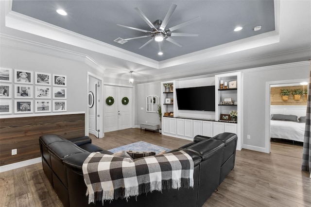 living room featuring ceiling fan, a raised ceiling, wood-type flooring, ornamental molding, and built in shelves