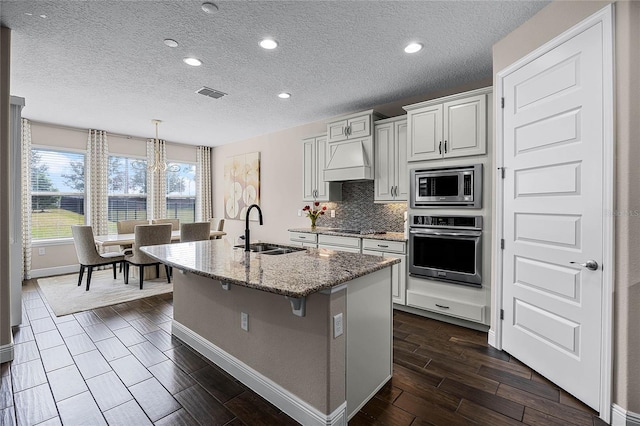 kitchen featuring decorative light fixtures, stainless steel appliances, premium range hood, sink, and a kitchen island with sink