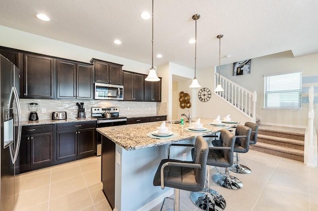 kitchen with stainless steel appliances, hanging light fixtures, a kitchen island with sink, light tile patterned flooring, and a breakfast bar area