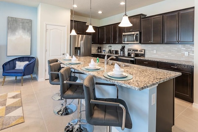 kitchen with a center island with sink, pendant lighting, light tile patterned flooring, stainless steel appliances, and light stone counters