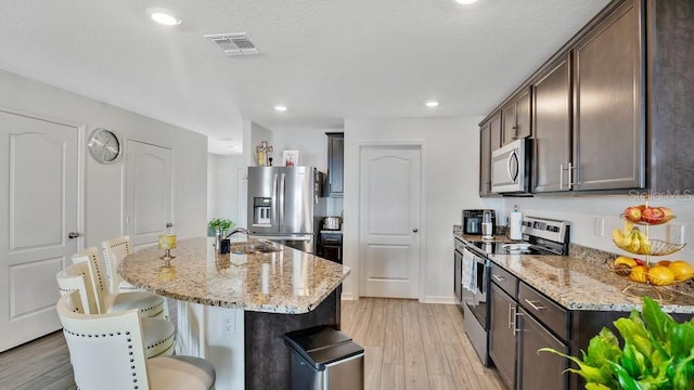 kitchen with sink, a kitchen island with sink, dark brown cabinetry, stainless steel appliances, and light stone countertops