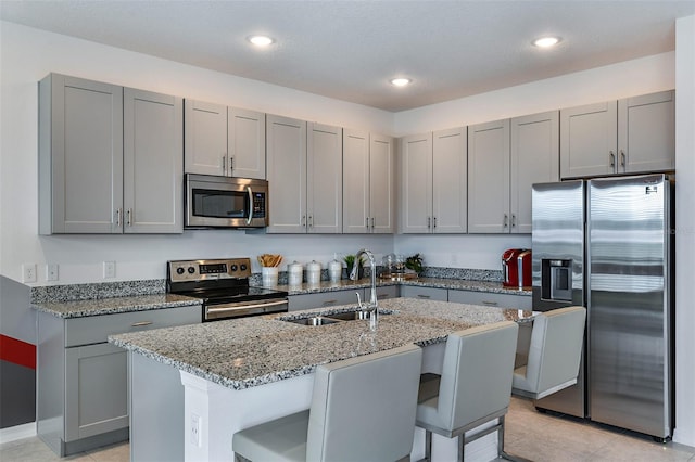 kitchen featuring a kitchen bar, sink, a kitchen island with sink, stainless steel appliances, and light stone counters