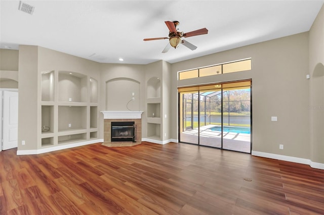 unfurnished living room featuring ceiling fan, a tiled fireplace, wood-type flooring, and built in shelves