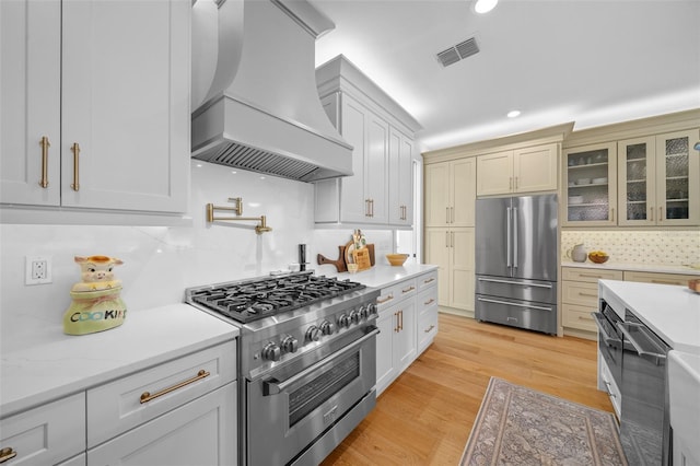 kitchen featuring high end appliances, tasteful backsplash, light wood-type flooring, custom range hood, and light stone countertops