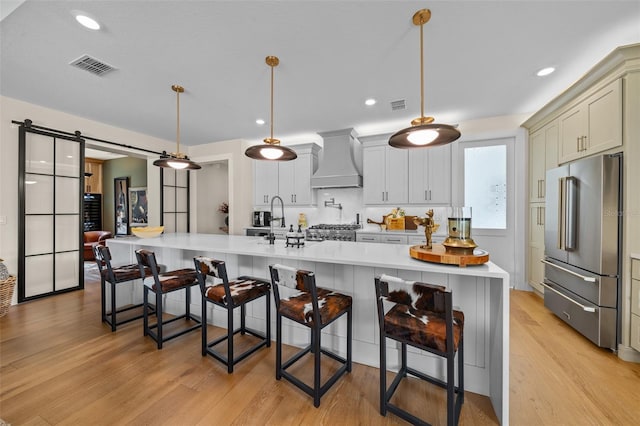 kitchen featuring a large island with sink, high quality fridge, pendant lighting, a barn door, and wall chimney range hood