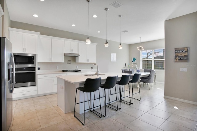 kitchen with appliances with stainless steel finishes, white cabinetry, a kitchen island with sink, light tile patterned floors, and decorative light fixtures