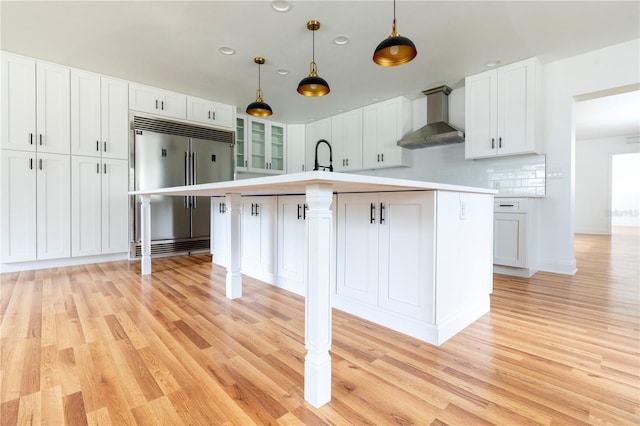 kitchen with stainless steel built in refrigerator, decorative light fixtures, an island with sink, wall chimney range hood, and white cabinets