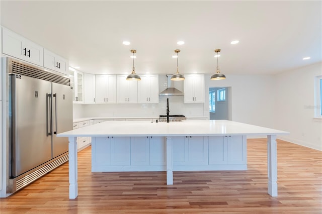 kitchen featuring decorative light fixtures, white cabinets, built in refrigerator, wall chimney range hood, and light wood-type flooring