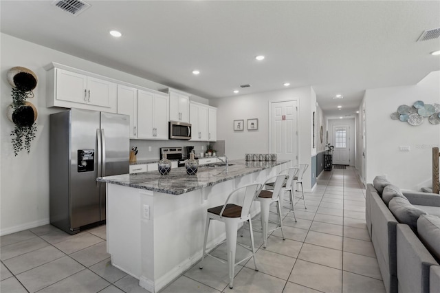 kitchen featuring white cabinetry, stainless steel appliances, dark stone countertops, an island with sink, and a kitchen breakfast bar