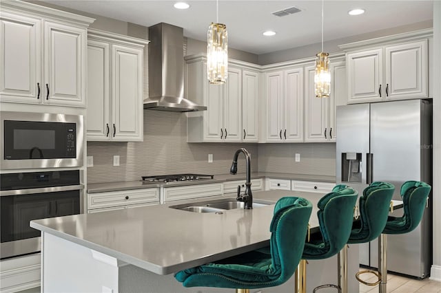 kitchen with sink, wall chimney range hood, an island with sink, stainless steel appliances, and white cabinets