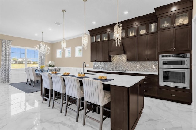 kitchen with crown molding, black electric stovetop, stainless steel double oven, decorative light fixtures, and an island with sink