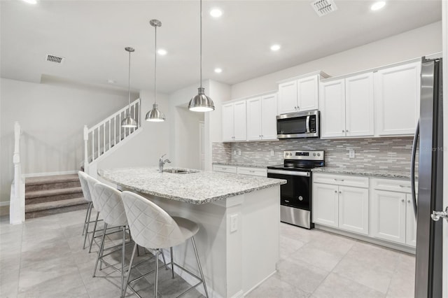 kitchen featuring appliances with stainless steel finishes, white cabinetry, light stone counters, a center island with sink, and decorative light fixtures