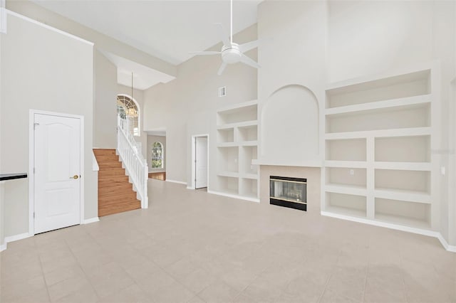 unfurnished living room with a tiled fireplace, a high ceiling, built in shelves, light tile patterned flooring, and ceiling fan with notable chandelier