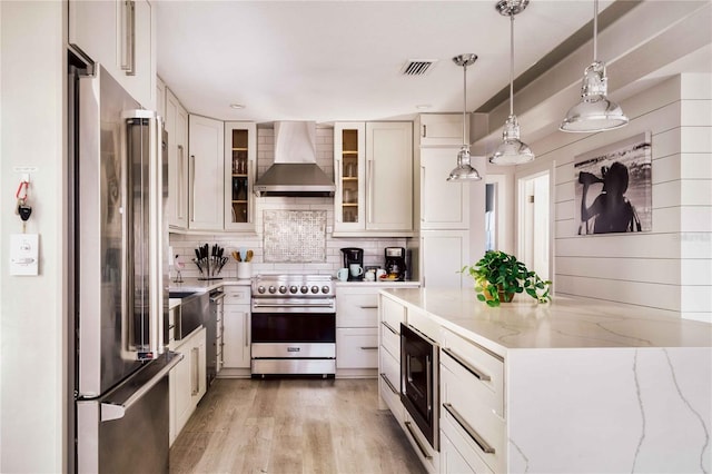 kitchen featuring pendant lighting, white cabinets, light stone counters, stainless steel appliances, and wall chimney range hood