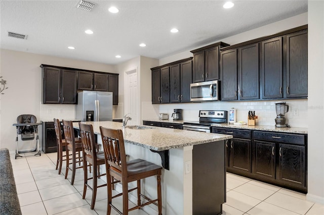 kitchen featuring a center island with sink, appliances with stainless steel finishes, light tile patterned flooring, light stone counters, and sink