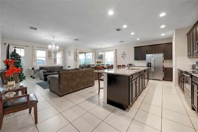 kitchen with sink, a kitchen island with sink, stainless steel appliances, light stone counters, and a chandelier