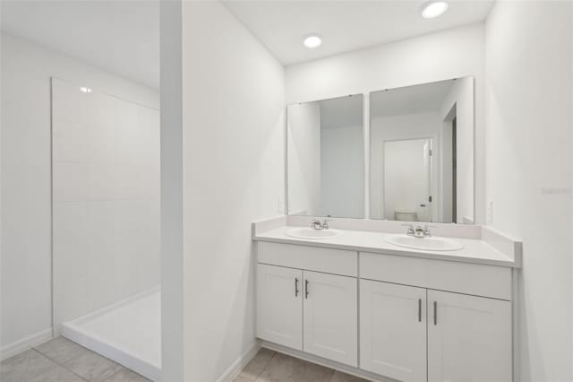 full bathroom with a tile shower, double vanity, a sink, and baseboards