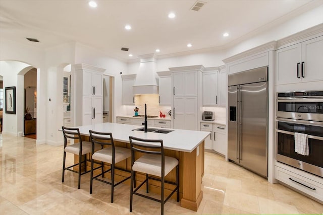 kitchen featuring a kitchen bar, white cabinetry, custom range hood, a kitchen island with sink, and stainless steel appliances