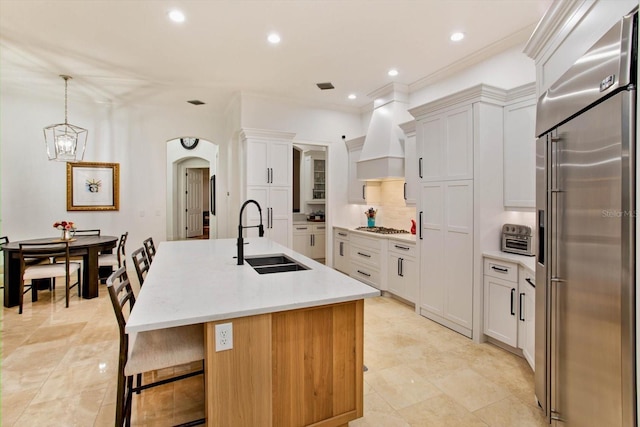 kitchen with pendant lighting, appliances with stainless steel finishes, custom exhaust hood, sink, and a center island with sink