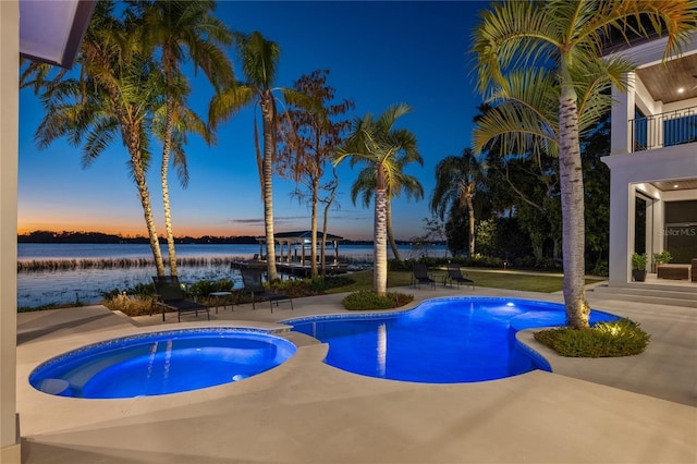 pool at dusk with an in ground hot tub, a patio, and a water view