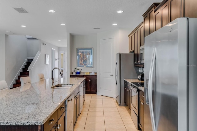 kitchen with sink, light tile patterned floors, appliances with stainless steel finishes, a kitchen island with sink, and light stone counters